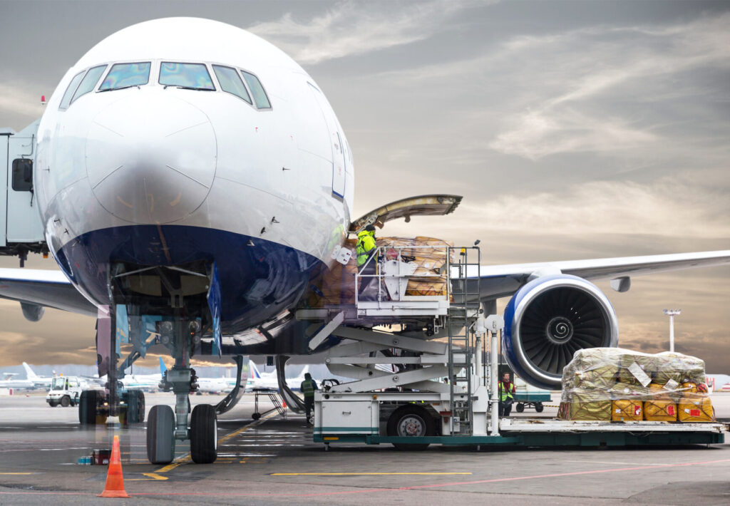 Aircraft being loaded with cargo by workers using a loader at the airport, with cloudy skies in the background.