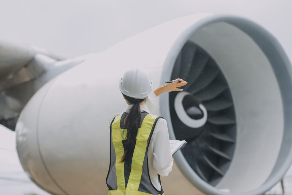 A person in a hard hat and safety vest inspects a large aircraft engine, holding a clipboard and pen.